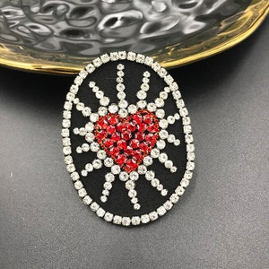 May include: A black oval-shaped patch with a red rhinestone heart surrounded by a circle of clear rhinestones.