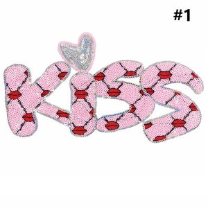 May include: A pink sequined patch spelling "KISS" with red lip accents and a silver heart. The letters are outlined in silver and have a diamond pattern. The patch is labeled with the number "#1" in the upper right corner.