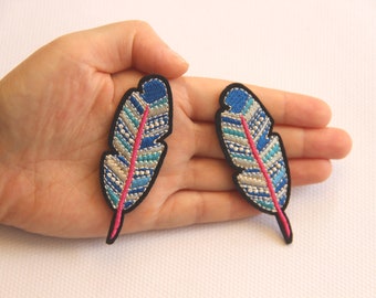 May include: Two embroidered feather patches. Each patch features a blue top, a pink stem, and a design of white, teal, and grey. The patches are outlined in black.