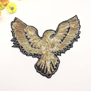 Può includere: Una toppa a forma di aquila dorata con paillettes e ali spiegate. La toppa ha un bordo nero ed è riempita di paillettes dorate, creando un effetto scintillante. Il design dell'aquila è dettagliato e adatto per abbellire abiti o accessori.