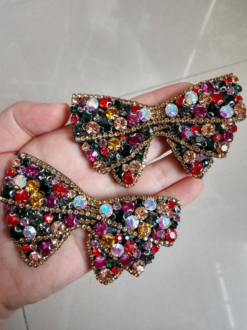 May include: Two decorative bow-shaped embellishments. Each bow is adorned with an array of colourful rhinestones in shades of red, green, yellow, and iridescent hues. The bows have a gold-coloured border.