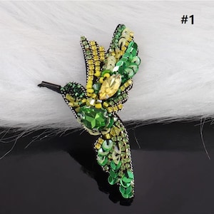 Puede incluir: Una aplicación de colibrí verde y amarilla con lentejuelas y cuentas. La aplicación está sobre un fondo blanco peludo. #1