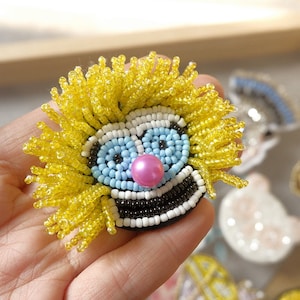 May include: A beaded clown face with yellow hair, blue eyes, a pink nose, and a black and white smile. The face is made of small beads and is attached to a fabric backing.