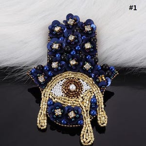May include: A blue and gold beaded hamsa hand with an evil eye charm. The hamsa is decorated with blue and gold beads and sequins.