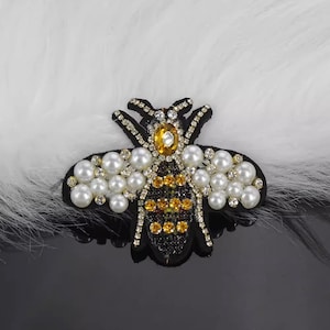 May include: A black and yellow bee embroidered patch with pearl accents. The bee is surrounded by white pearls and has a clear rhinestone in the center of its body.