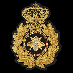 May include: A gold embroidered patch featuring a bee design encircled by a wreath. The bee is embellished with rhinestones and the wreath is crafted from gold thread. A gold crown sits atop the wreath.