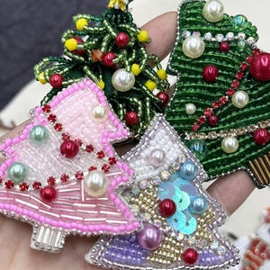 Könnte beinhalten: Nahaufnahme von vier weihnachtsbaumförmigen Broschen. Die Broschen sind mit Perlen, Pailletten und Kunstperlen in verschiedenen Farben verziert, darunter Grün, Rosa und Lila. Die Broschen sind aufwendig detailliert und festlich.