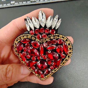 May include: A red heart-shaped brooch with a crown of clear rhinestones and a border of gold beads. The heart is covered in red rhinestones of various sizes.