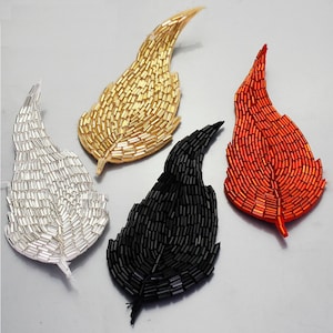 May include: Four beaded leaf appliques in silver, gold, black, and orange. Each leaf is intricately detailed with small beads, creating a textured surface. The leaves are arranged on a light grey surface, showcasing their vibrant colours and detailed craftsmanship.
