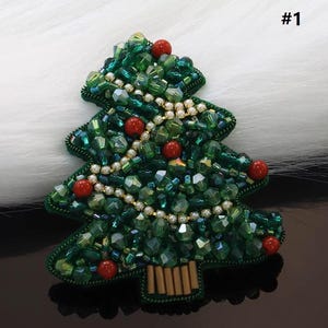Puede incluir: Un broche de árbol de Navidad, intrincadamente bordado con cristales verdes e iridiscentes, cuentas rojas y pequeñas perlas blancas. El broche tiene un tronco marrón y está sobre un fondo blanco. El número "#1" está en la esquina superior derecha.