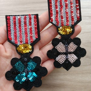 Könnte beinhalten: Zwei gestickte Patches mit einem Schmetterlingsmotiv. Ein Patch ist schwarz mit einem türkisfarbenen Schmetterling und der andere ist schwarz mit einem rosa Schmetterling. Beide Patches haben goldene Pailletten an den Rändern.