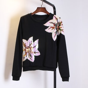 May include: Black long-sleeve sweatshirt with two large sequined flower designs. The flowers feature pink, white, and gold sequins. The sweatshirt is hanging on a black clothing rack.