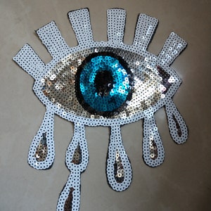 May include: A white and silver sequin eye with a blue sequin pupil and six white sequin teardrop shapes hanging below.