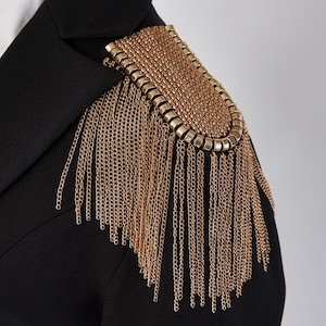 Peut inclure: Un blazer noir avec une épaulette en chaîne dorée. Le détail de l'épaule présente un motif rectangulaire avec des rangées de petits maillons dorés et des chaînes pendantes. Le tissu du blazer est lisse et sombre.