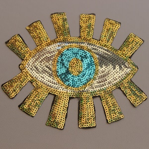 May include: A sequined eye patch with a turquoise iris and a gold and black sunburst design. The eye is detailed with silver and white sequins. The patch is likely for clothing or accessories.