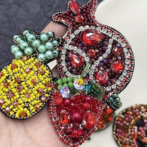 Puede incluir: Primer plano de tres broches de frutas con cuentas: una piña amarilla, una granada roja y una fresa roja. Los broches están adornados con cuentas de colores y pedrería brillante, creando un diseño lujoso y detallado. Los broches están colocados sobre un fondo neutro.