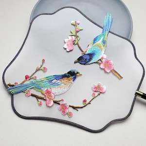 May include: Embroidered fan featuring two colorful birds perched on flowering branches. The birds have blue, green, and yellow plumage, with pink blossoms and brown branches. The fan has a gray background and a dark gray border.