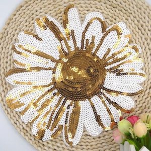 May include: A large, sequined daisy applique. The flower features white petals with gold accents and a gold center. The applique is likely for clothing or craft projects. The flower is on a woven, beige surface.
