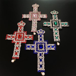 Puede incluir: Cuatro colgantes de cruz enjoyados en rojo, rosa, azul y verde. Cada cruz está adornada con diamantes de imitación y una gota de perla.