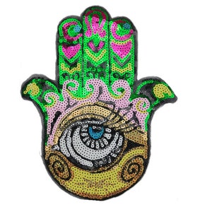 Può includere: Una mano di hamsa verde, rosa e dorata con un occhio blu al centro. La hamsa è fatta di paillettes ed è un simbolo popolare di protezione e buona fortuna.