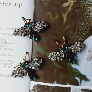 Puede incluir: Tres broches en forma de abeja de color verde y plateado con pedrería y detalles en verde esmeralda. Los broches están sobre una superficie con estampado marrón y blanco.