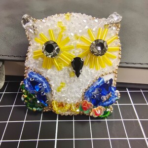 May include: A white owl brooch with yellow and blue bead embellishments. The owl's eyes are black and white beads. The brooch is made of beads and sequins.