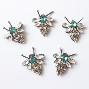 May include: Five bee-shaped embellishments with clear and green rhinestones. The bees have black wire legs and a black body.