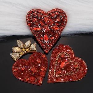 Puede incluir: Un surtido de broches rojos en forma de corazón y una pequeña corona, todos adornados con brillantes cristales rojos y transparentes. Los broches varían en diseño, con intrincados trabajos de abalorios y arreglos de gemas. La corona es dorada con cristales transparentes.
