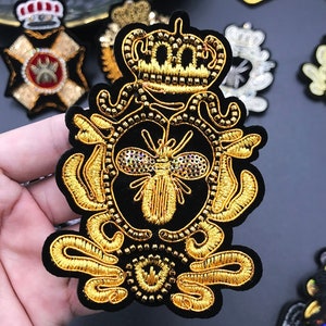 May include: A gold embroidered patch featuring a crown, a bee with rhinestones, and intricate details on a black velvet background.
