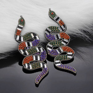 Puede incluir: Un par de pendientes con forma de serpiente. Los pendientes están adornados con pedrería de colores rojo, verde, azul y blanco, con detalles en negro. Los pendientes están sobre una superficie negra reflectante con un fondo blanco y esponjoso.