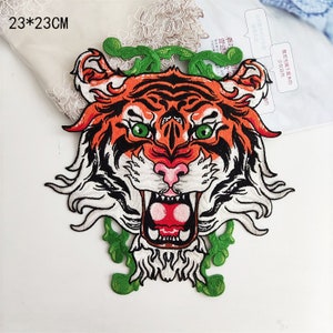 Può includere: Un grande toppa ricamata da stirare con il viso di una tigre con occhi verdi e bocca aperta. La tigre è circondata da foglie verdi. La toppa misura 23 cm per 23 cm.