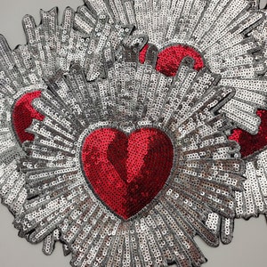 以下が含まれることがあります： ハート型のスパンコールパッチのクローズアップ。各パッチは、中心に鮮やかな赤いハートがあり、そこから外側に広がる銀色のスパンコールで囲まれています。パッチは黒い縁取りがあり、明るい灰色の表面に配置されています。
