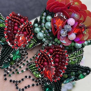 Può includere: Primo piano di una spilla floreale fatta a mano. La spilla presenta petali di paillettes rosse, gemme color rubino e perline verdi. Il design incorpora foglie verdi e dettagli di perline nere, creando un accessorio lussuoso e dettagliato.
