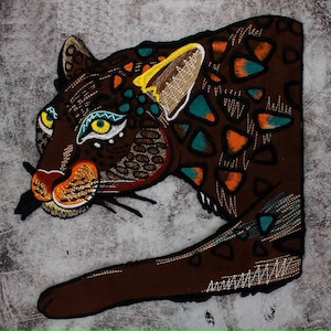 May include: Embroidered patch of a brown jaguar with teal and orange markings. The jaguar's eyes are green and yellow. The patch is on a grey background.