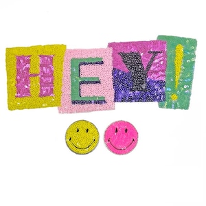 Könnte beinhalten: Ein Set aus bunten, mit Pailletten besetzten Patches, die "HEY!" buchstabieren, mit zwei Smiley-Patches darunter. Die Patches sind rosa, gelb, grün und lila.