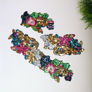 May include: Four floral sequin appliques in various sizes. The appliques feature a mix of colors, including gold, pink, green, and blue sequins, arranged in a floral pattern. Each applique has a black backing. The appliques are ideal for embellishing clothing or accessories.