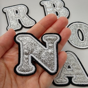 Puede incluir: Letras de planchar de purpurina plateada en un estilo de fuente en negrita. Las letras están delineadas en blanco y negro. Las letras son N, R, O, B y A.