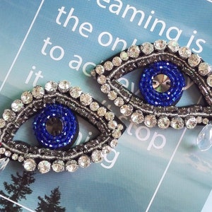 May include: Two blue evil eye charms with crystal embellishments and a teardrop crystal dangle. The charms are beaded with blue and silver accents.