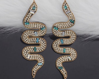 May include: A pair of long, dangling earrings shaped like snakes. The earrings are made of gold-toned metal and are covered in clear and green rhinestones.