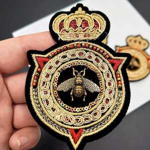 May include: Embroidered patch with a gold bee at the centre, encircled by gold and red detailing, and a gold crown. The patch is set on black velvet. The design is intricate and detailed, with a vintage aesthetic.