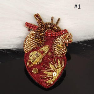 Può includere: Una spilla a forma di cuore realizzata in tessuto rosso scuro, decorata con perline, paillettes e cristalli dorati, bronzo e rossi. Il design presenta un pianeta dorato ricamato con anelli, piccole paillettes a forma di stella e fili dorati.
