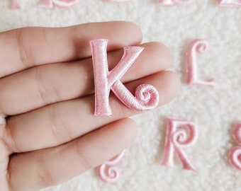 May include: Pink felt letter K with a swirl at the end of the letter. The letter is held in a hand.