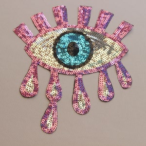 May include: A pink and blue sequin eye patch with a teardrop design. The eye is blue with a black pupil and a gold iris.