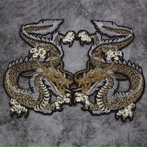 Puede incluir: Dos parches de dragón bordados en oro y plata con lentejuelas. Los dragones se miran, con la cabeza echada hacia atrás y la boca abierta.