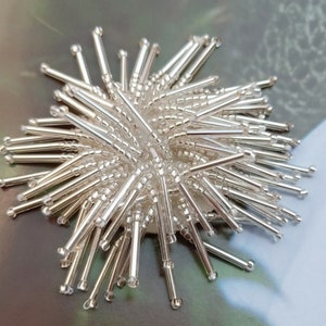 May include: A close-up of a silver beaded embellishment. The beads are arranged in a circular shape, creating a spiky, starburst effect.
