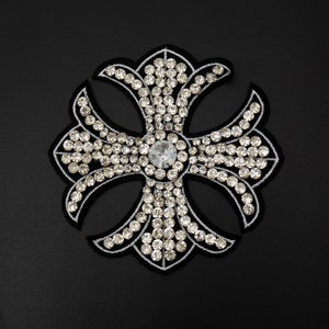 May include: A black and white cross-shaped patch with a large clear rhinestone in the centre. The cross is covered in smaller rhinestones.