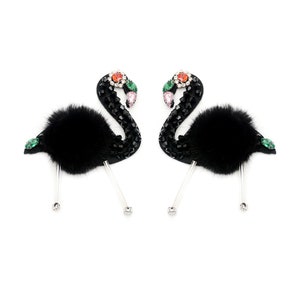 Puede incluir: Un par de pendientes negros con forma de flamenco. Los pendientes presentan cuerpos de piel negra, cuellos adornados con cuentas negras y acentos de joyas de colores en la cabeza. Tienen postes transparentes con una pequeña joya redonda en el extremo.