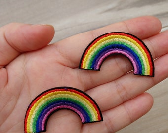 May include: Two rainbow-coloured embroidered patches in the shape of an arch. The patches are on a hand.