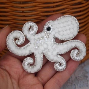 Könnte beinhalten: Weißer Oktopus-Broschen mit Perlenbesatz. Der Oktopus hat acht sich kräuselnde Arme und ein schwarzes Auge. Die Brosche besteht aus weißen Perlen und ist mit silbernen Perlen umrandet. Der Hintergrund ist ein geflochtener Korb.