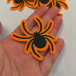 May include: A black and orange embroidered spider patch featuring a central circular body and eight curved legs. The design is outlined in bright orange thread with a solid black interior, creating a high-contrast graphic suitable for fabric application.